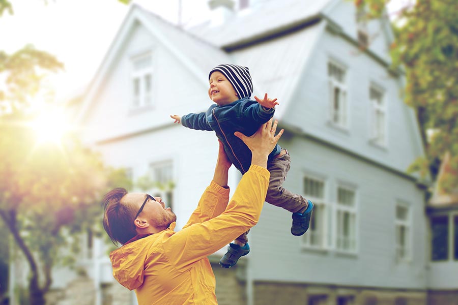 Personal Insurance - Father Lifts Toddler Son Into the Air in Front of Their Large White Home